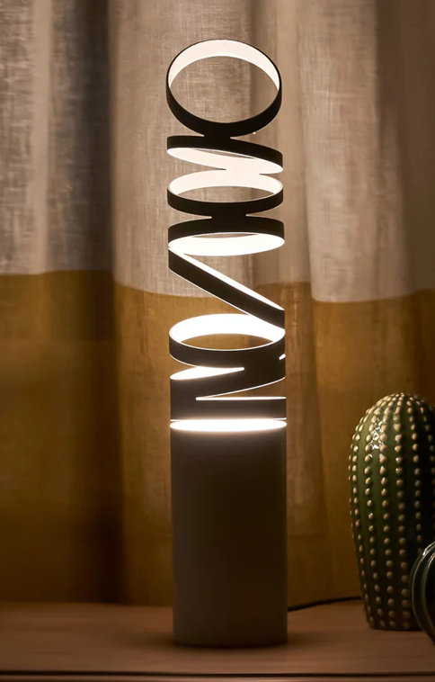 Whirl Table Lamp — spiral table light on a dark cylindrical base | أباجورة ويرل، تصميم حلزوني مضيء على قاعدة أسطوانية داكنة